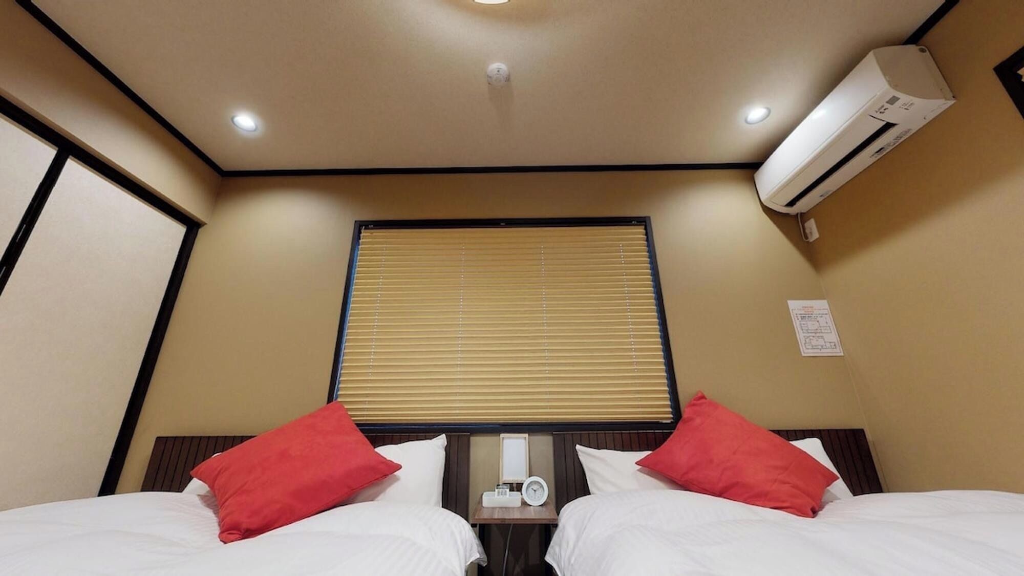 A hotel room with two beds, a window with blinds, an air conditioning unit, and a bedside table with a clock.