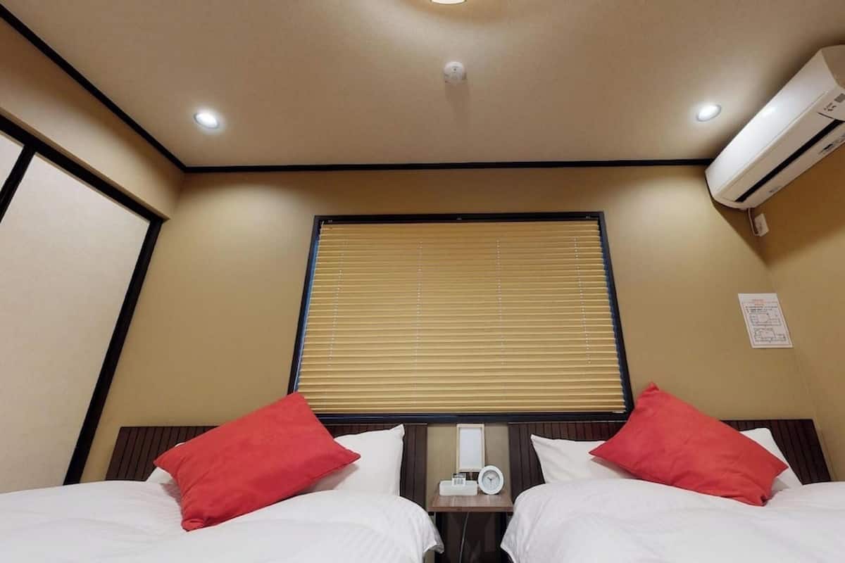 A hotel room with two beds, a window with blinds, an air conditioning unit, and a bedside table with a clock.