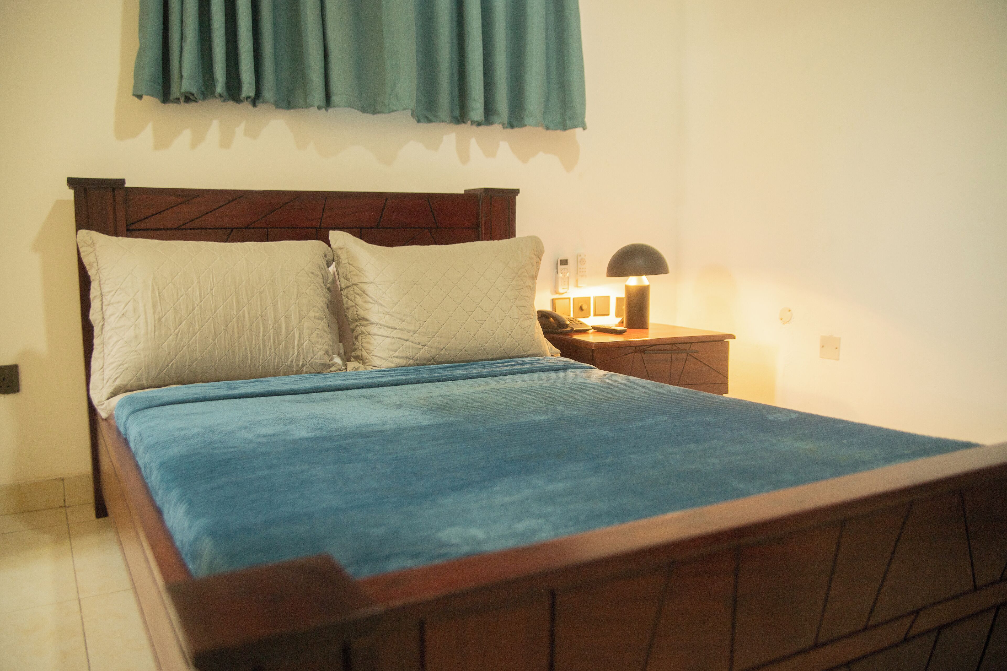 A neatly made bed with a blue blanket and two pillows, a wooden headboard, a bedside table with a lamp, and a wall-mounted curtain.