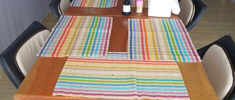 Un comedor con una mesa de madera, individuales a rayas y sillas acolchadas. Se ve un refrigerador con bebidas al fondo.