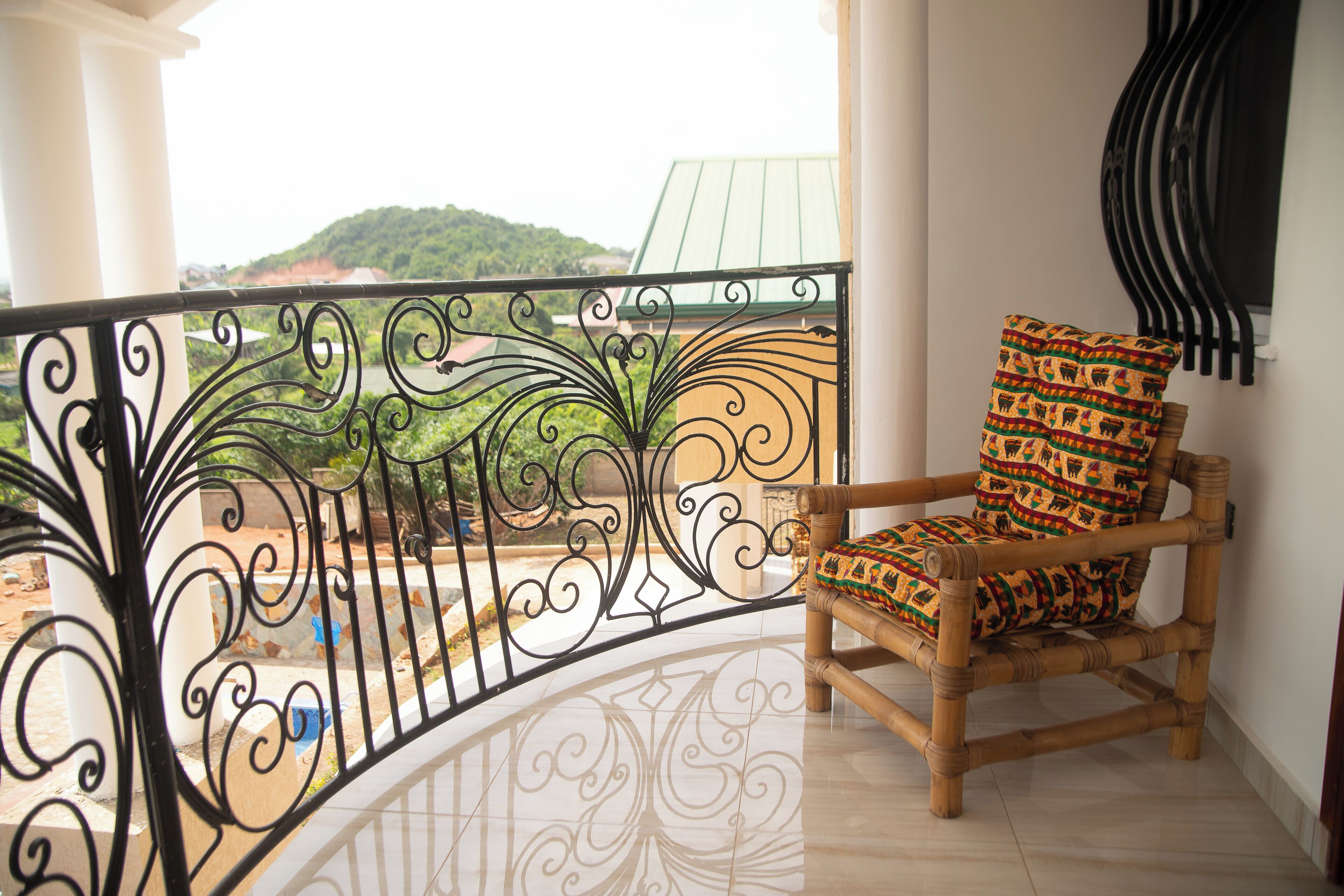 A balcony with a wrought iron railing, a bamboo chair with patterned cushions, and a view of a green landscape.