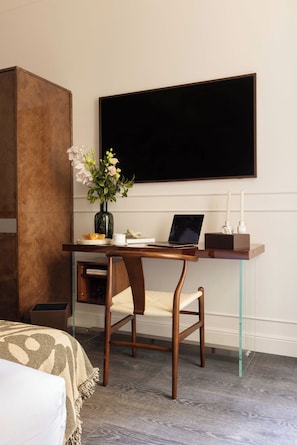 Une chambre d’hôtel avec un bureau en bois, un ordinateur portable, un vase avec des fleurs, un lit recouvert d’une couverture à motifs et un grand écran noir fixé au mur.