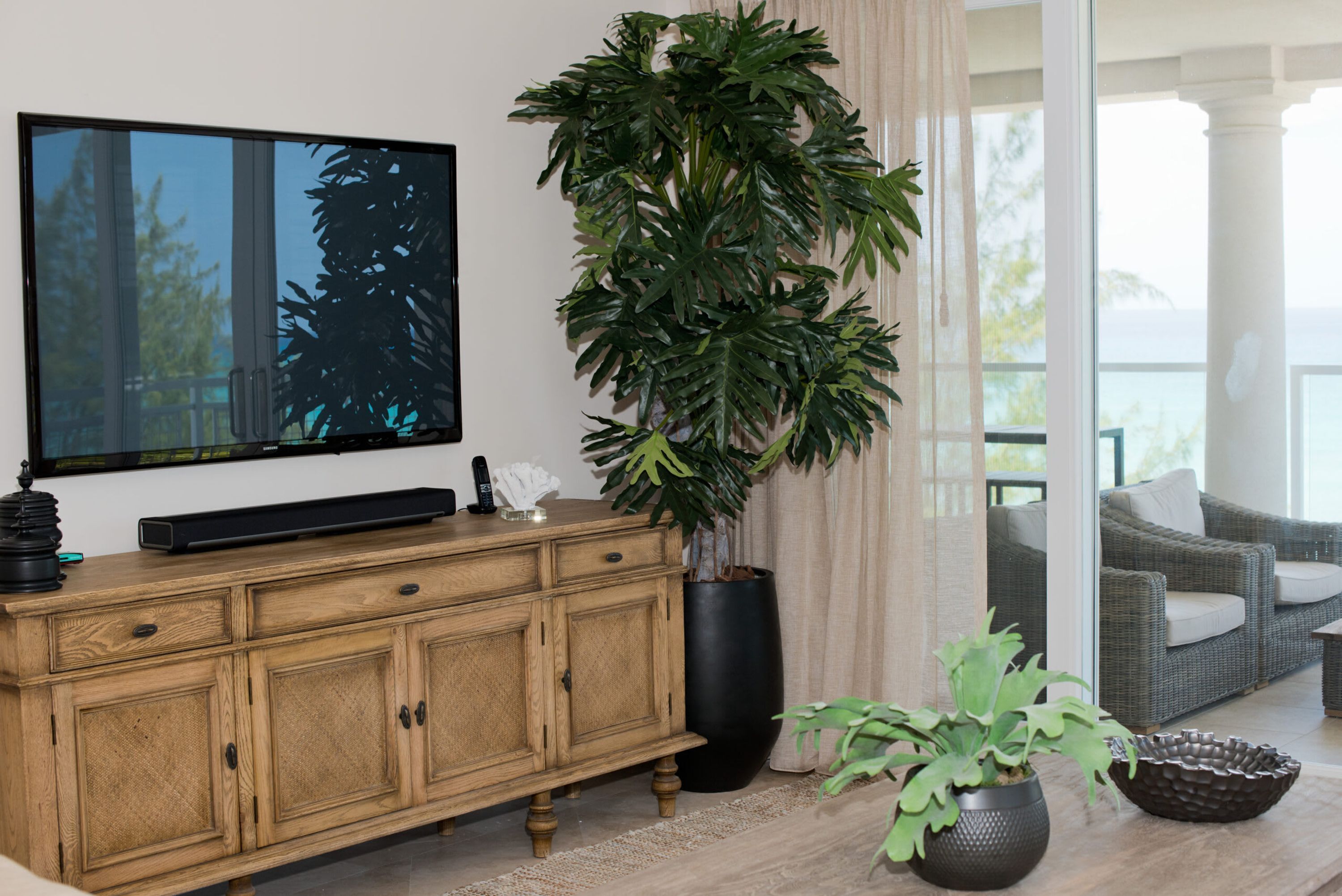A living room with a flat-screen TV, wooden cabinet, large potted plant, wicker sofa, and a view of the outdoors.