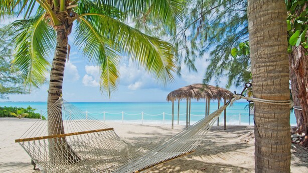 A hammock tied between two palm trees on a sandy beach with a thatched-roof structure in the background. - Seabreeze 8 - Luxury 4-bedroom on Seven Mile Beach (Seven Mile Beach)