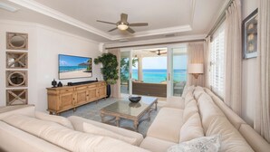 A spacious living room with a large sofa, a coffee table, and a television. There is a view of the ocean through large windows. - Seabreeze 8 - Luxury 4-bedroom on Seven Mile Beach (Seven Mile Beach)