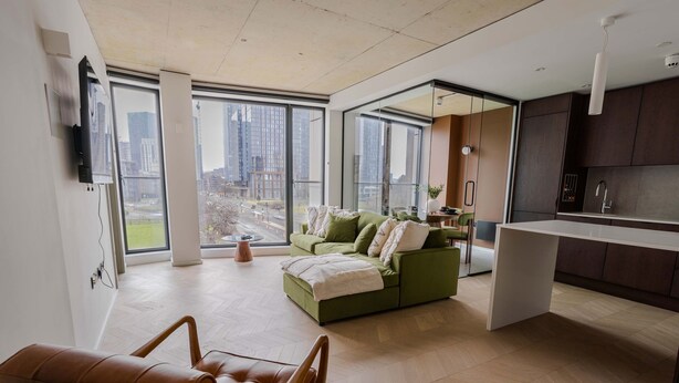 Una sala de estar moderna con un sofá verde, un sillón de cuero marrón y una cocina con armarios oscuros y encimera blanca.