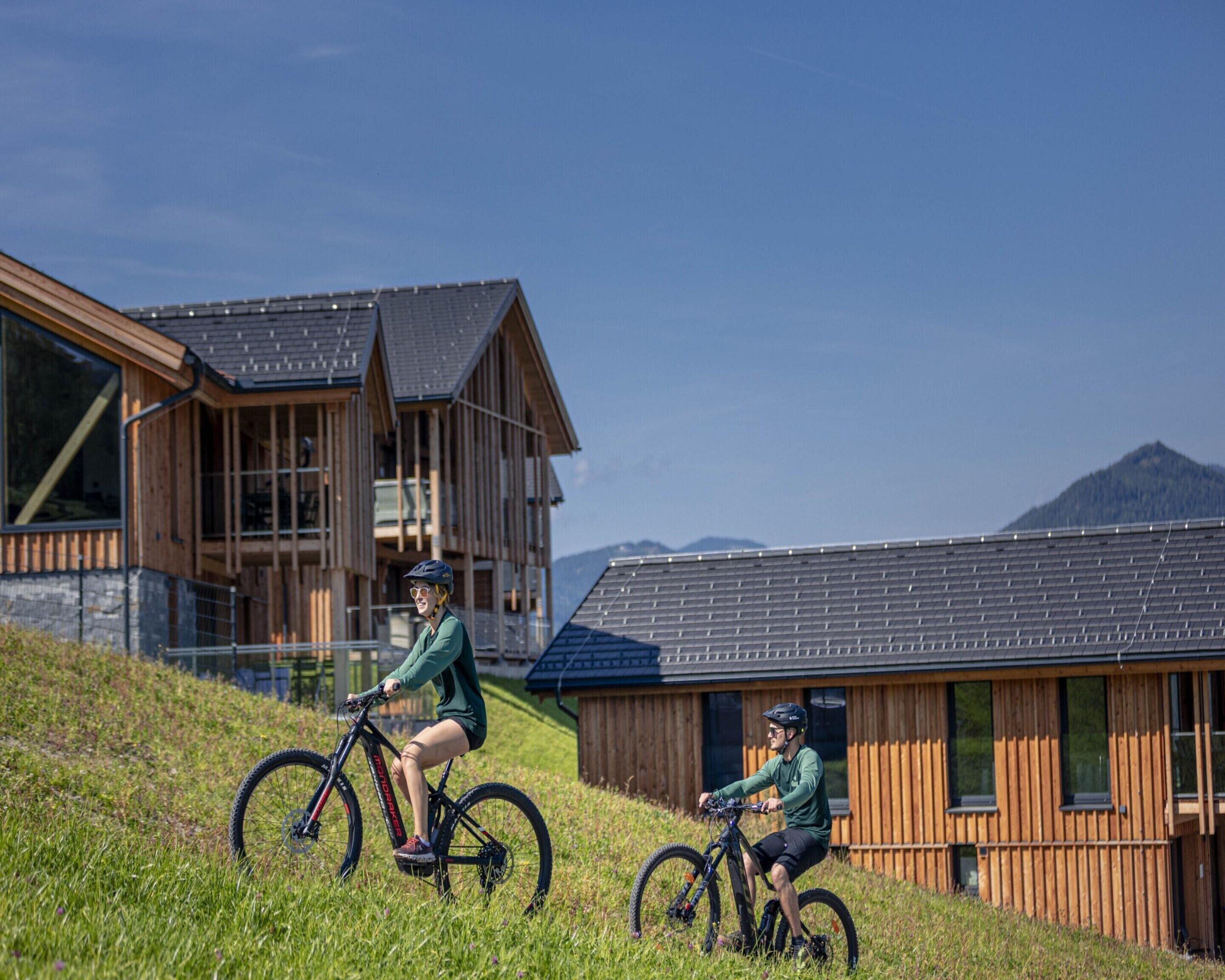 Deux personnes font du vélo sur une pente herbeuse, avec des bâtiments modernes en bois en arrière-plan.