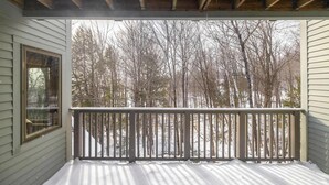 A covered porch with a wooden railing, a window, and a view of leafless trees and snow-covered ground.