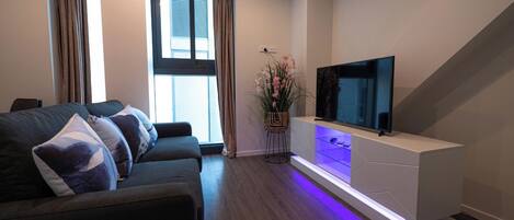 A modern living room with a dark-colored sofa, a flat-screen TV on a white entertainment unit, and a potted plant with pink flowers.