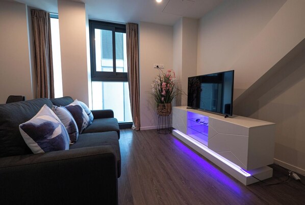 Un salon moderne avec un canapé de couleur foncée, un téléviseur à écran plat posé sur un meuble blanc, et une plante en pot ornée de fleurs roses.