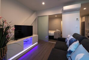 Un salon moderne avec un téléviseur à écran plat posé sur une console blanche, un canapé de couleur foncée agrémenté de coussins à motifs et une petite table d’appoint sur laquelle se trouve un vase de fleurs.