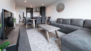 A modern living room with a grey sectional sofa, a wooden coffee table, and a dining area with grey chairs.