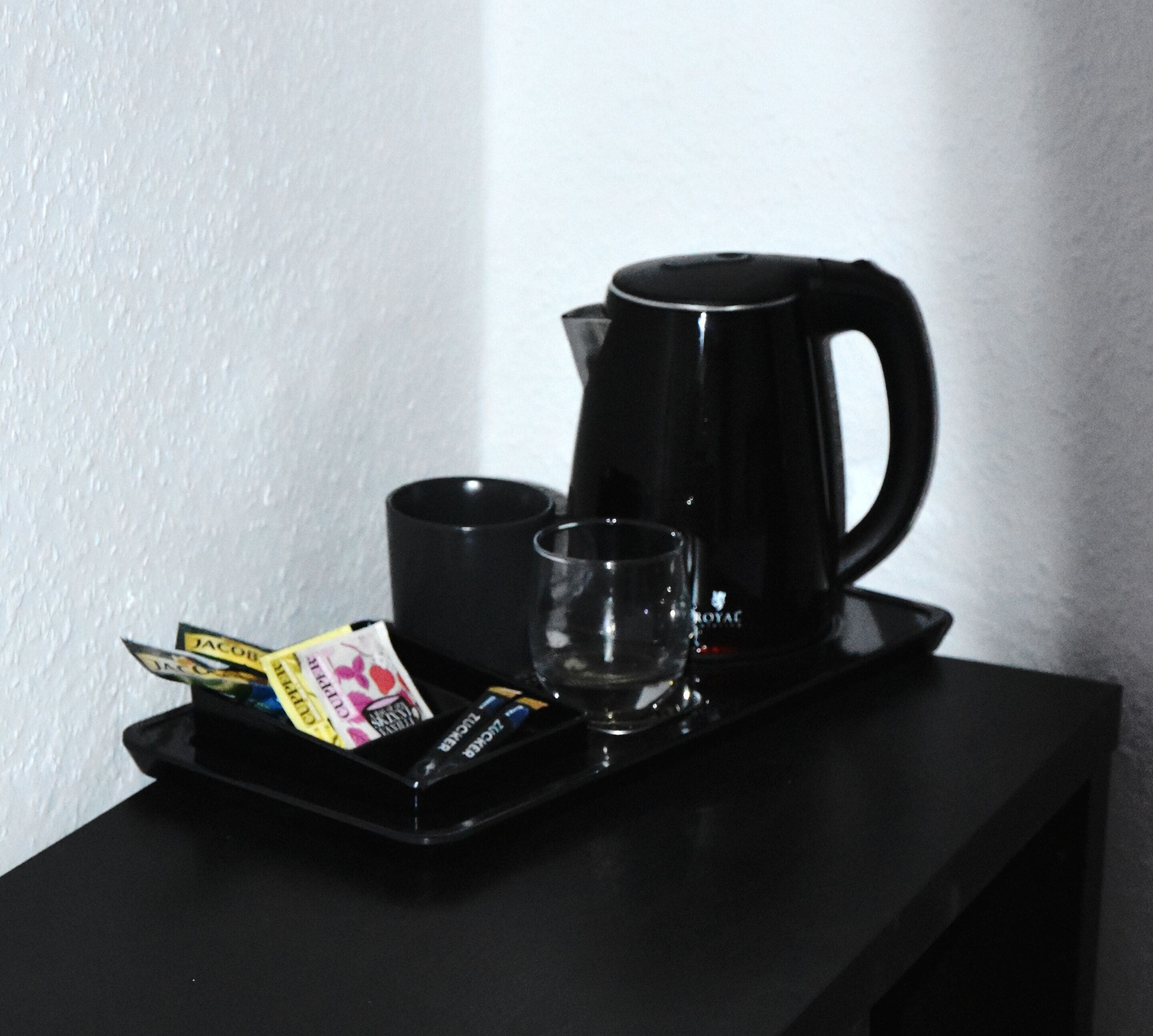 A black tray with a coffee maker, a cup, a glass, and some toiletries on a black table.