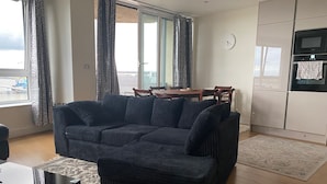 A living room with a sofa, a dining table, and chairs. There is a window with curtains, a clock on the wall, and a kitchen area with appliances. - 2 Bed Flat - Sleeps 4 - Parking - Wifi (London)