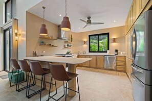 Une cuisine moderne dotée d’un îlot central, de tabourets de bar, de luminaires suspendus, d’un ventilateur de plafond et d’un grand réfrigérateur.