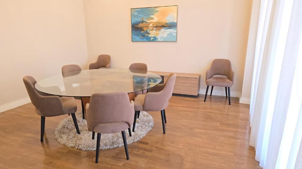 Una mesa redonda de cristal con seis sillas, una alfombra mullida, un cuadro en la pared y suelo de madera.