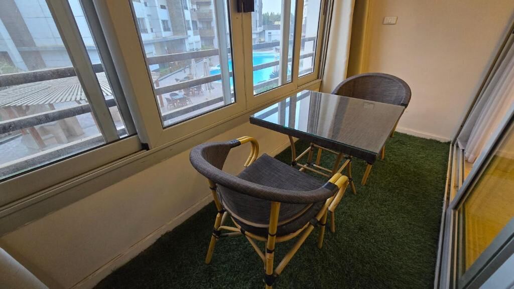 Une pièce avec une table en verre et deux chaises, de grandes fenêtres donnant sur des immeubles et une piscine, et une moquette verte.