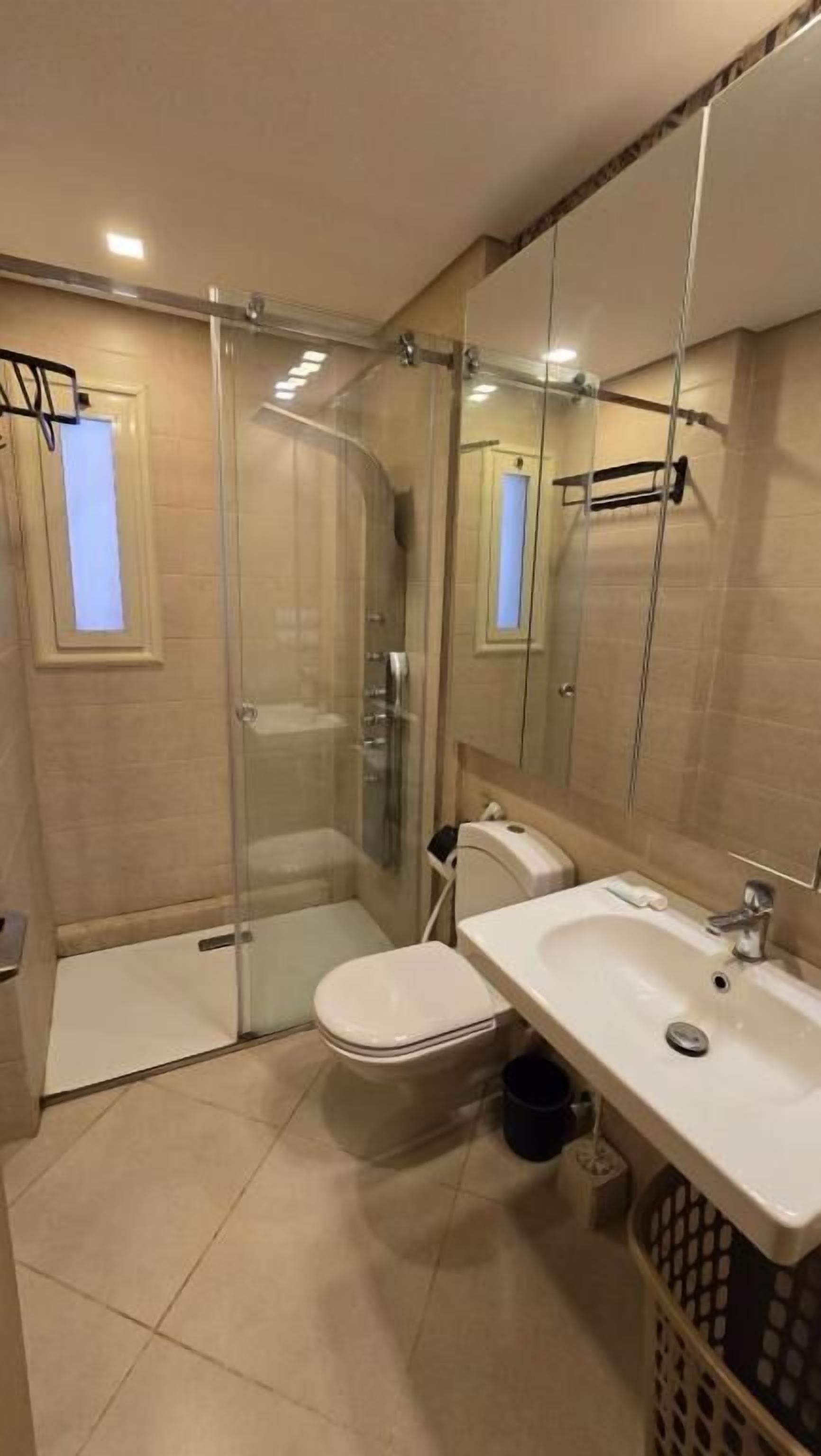 A modern bathroom with a glass-enclosed shower, a white toilet, a sink with a mirror, and a laundry basket.