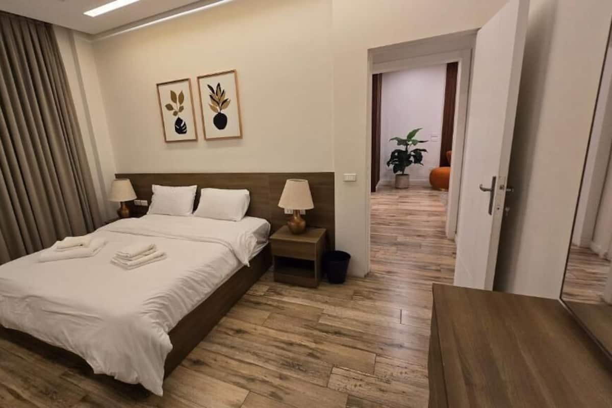 Une chambre d’hôtel avec un lit, des tables de chevet, une lampe, une plante en pot et une porte donnant sur une autre pièce.