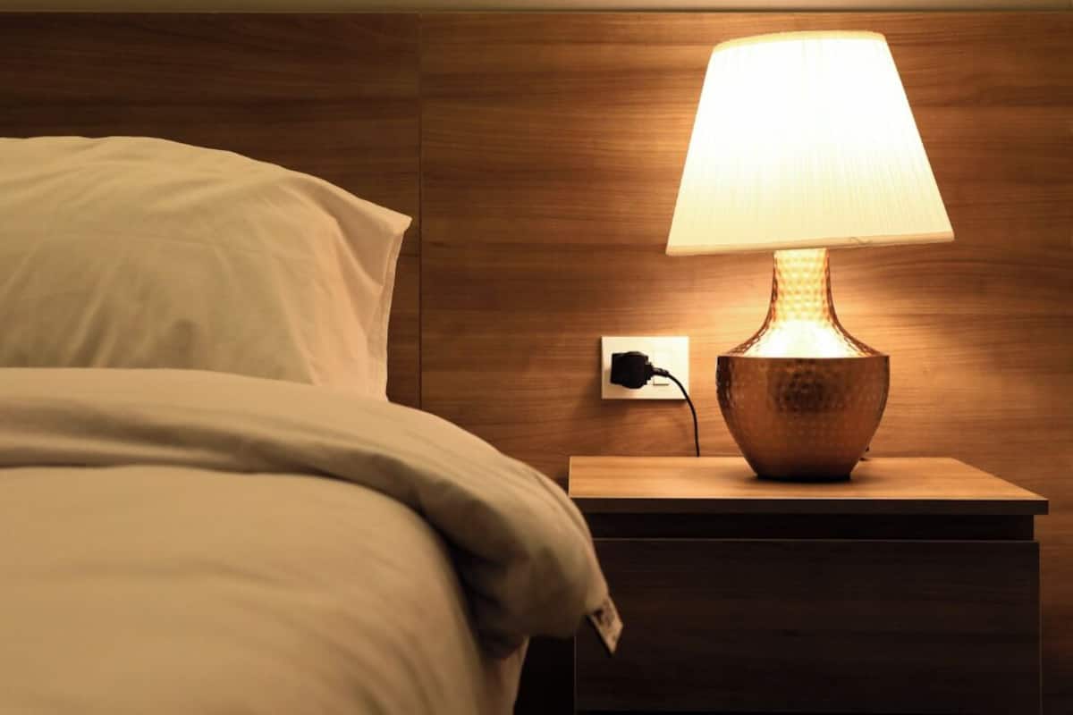 Un lit bien fait, avec une couette blanche et des oreillers, une table de chevet en bois avec une lampe, et une prise de courant sur le mur.