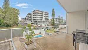 A balcony with a view of a multi-story building, a swimming pool, and a road with cars.