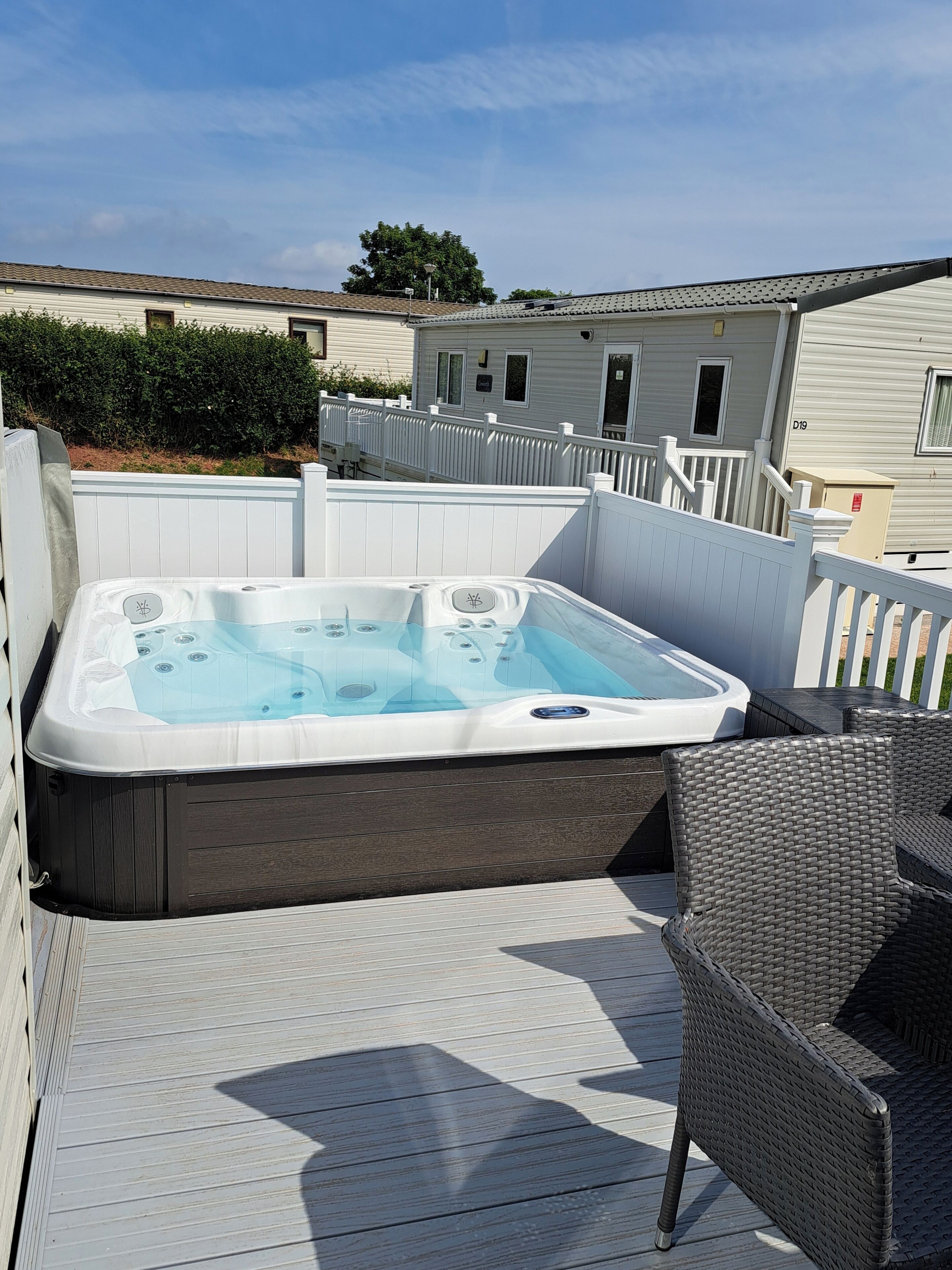 A hot tub on a wooden deck with wicker furniture, surrounded by white fences and a row of houses in the background.