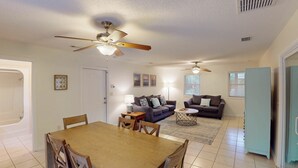 A living room with a sofa, coffee table, and ceiling fan. There is a dining table with chairs and a window with blinds.