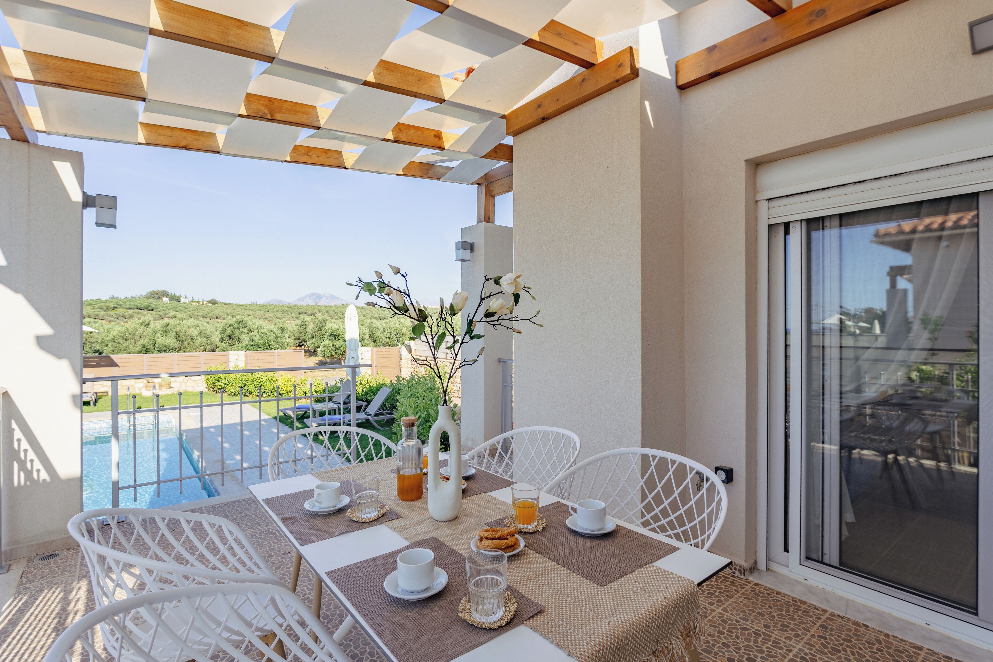 Un balcón con una mesa dispuesta para el desayuno, sillas blancas y vistas a una piscina y vegetación.