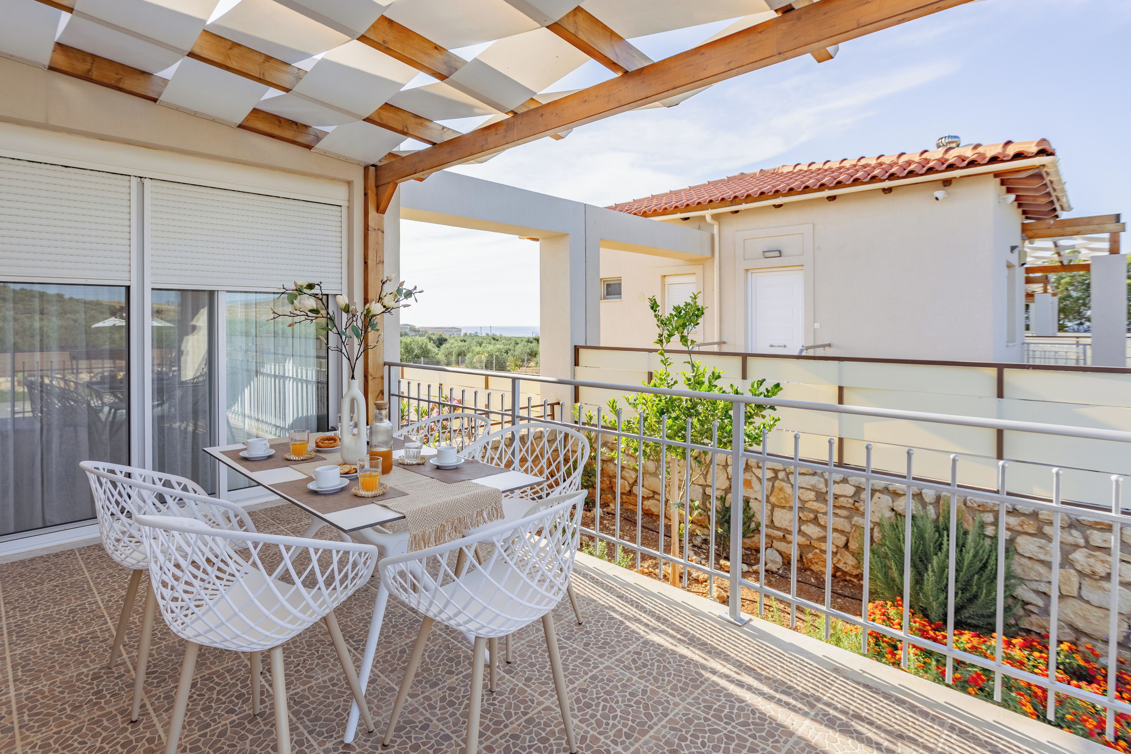 A balcony with white chairs, a table with a glass of orange juice, and a view of a stone wall and red flowers.