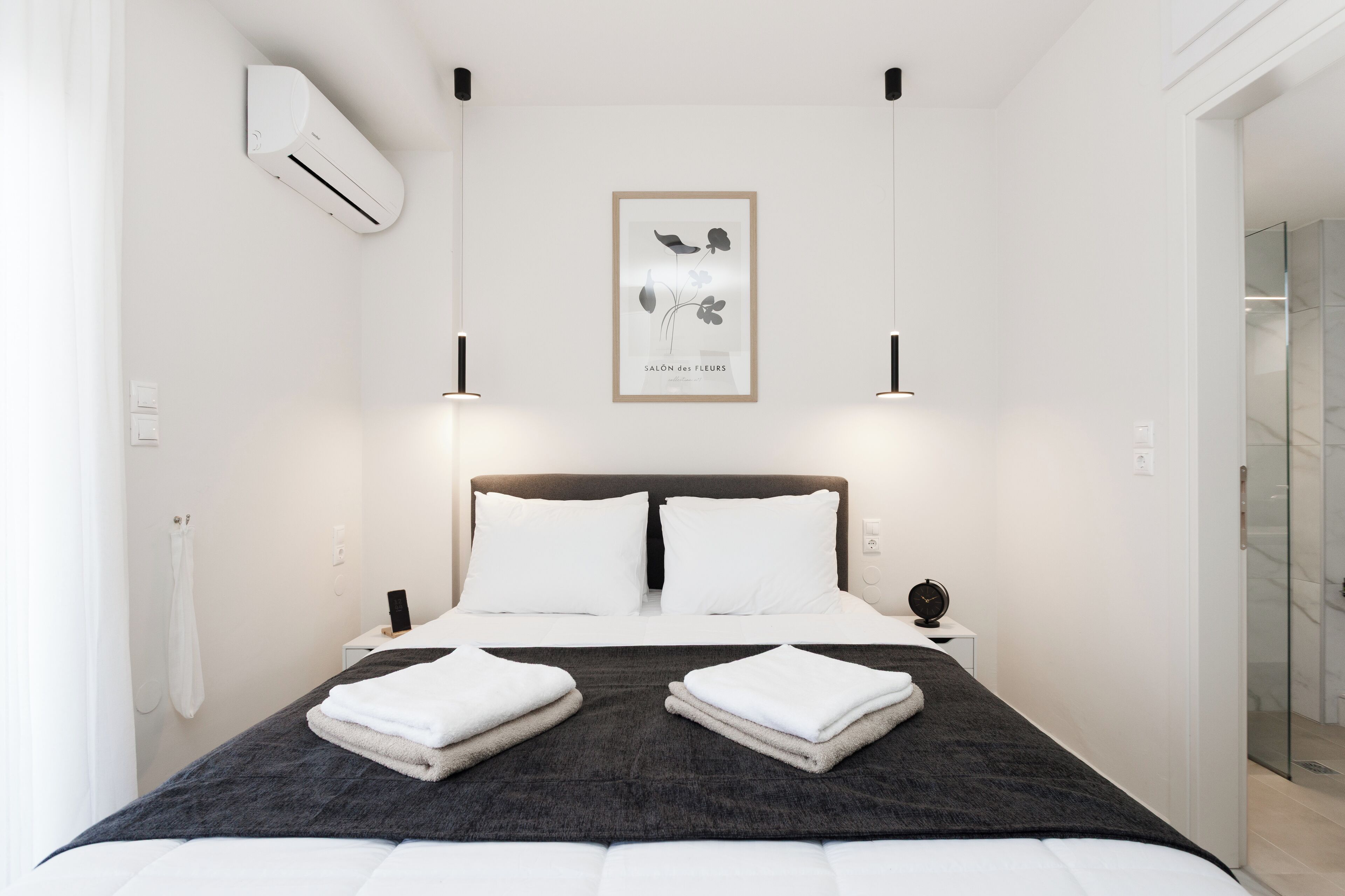 A modern hotel room with a large bed, two bedside tables, a wall-mounted air conditioner, and a framed botanical print on the wall.