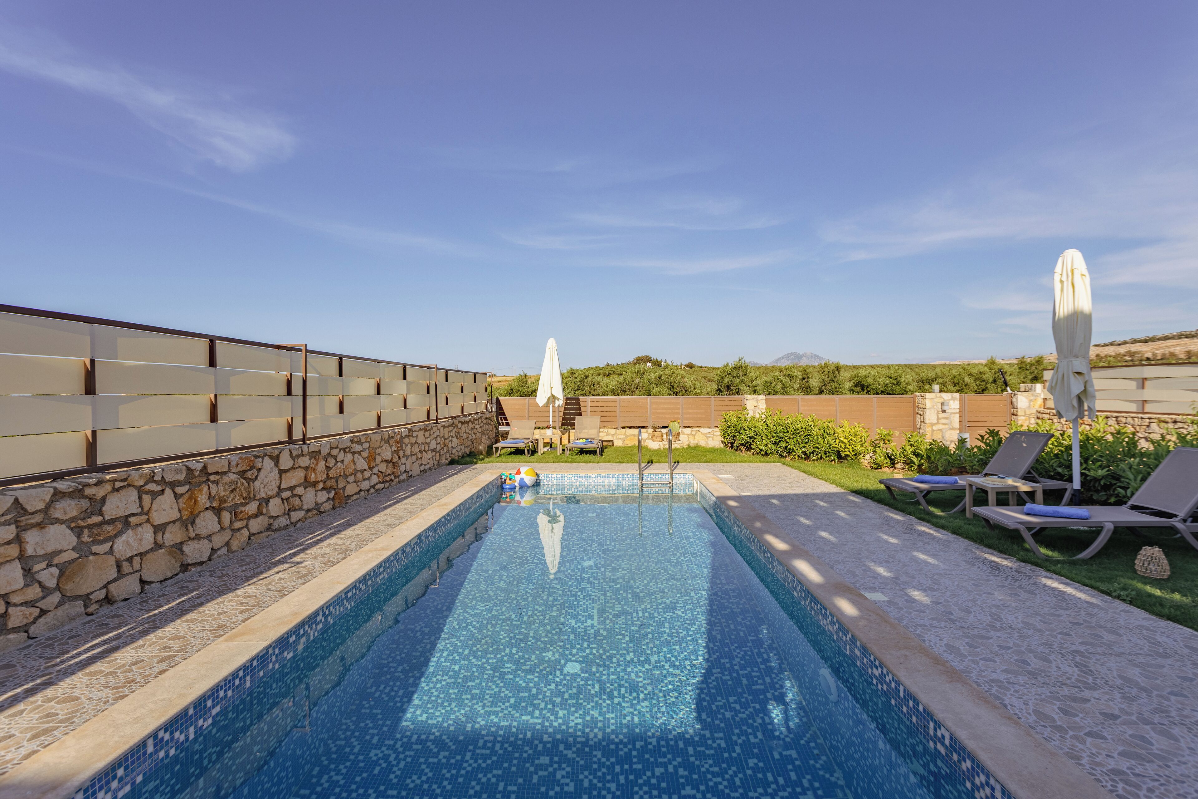 A swimming pool with lounge chairs and umbrellas, surrounded by a stone wall and a view of hills.
