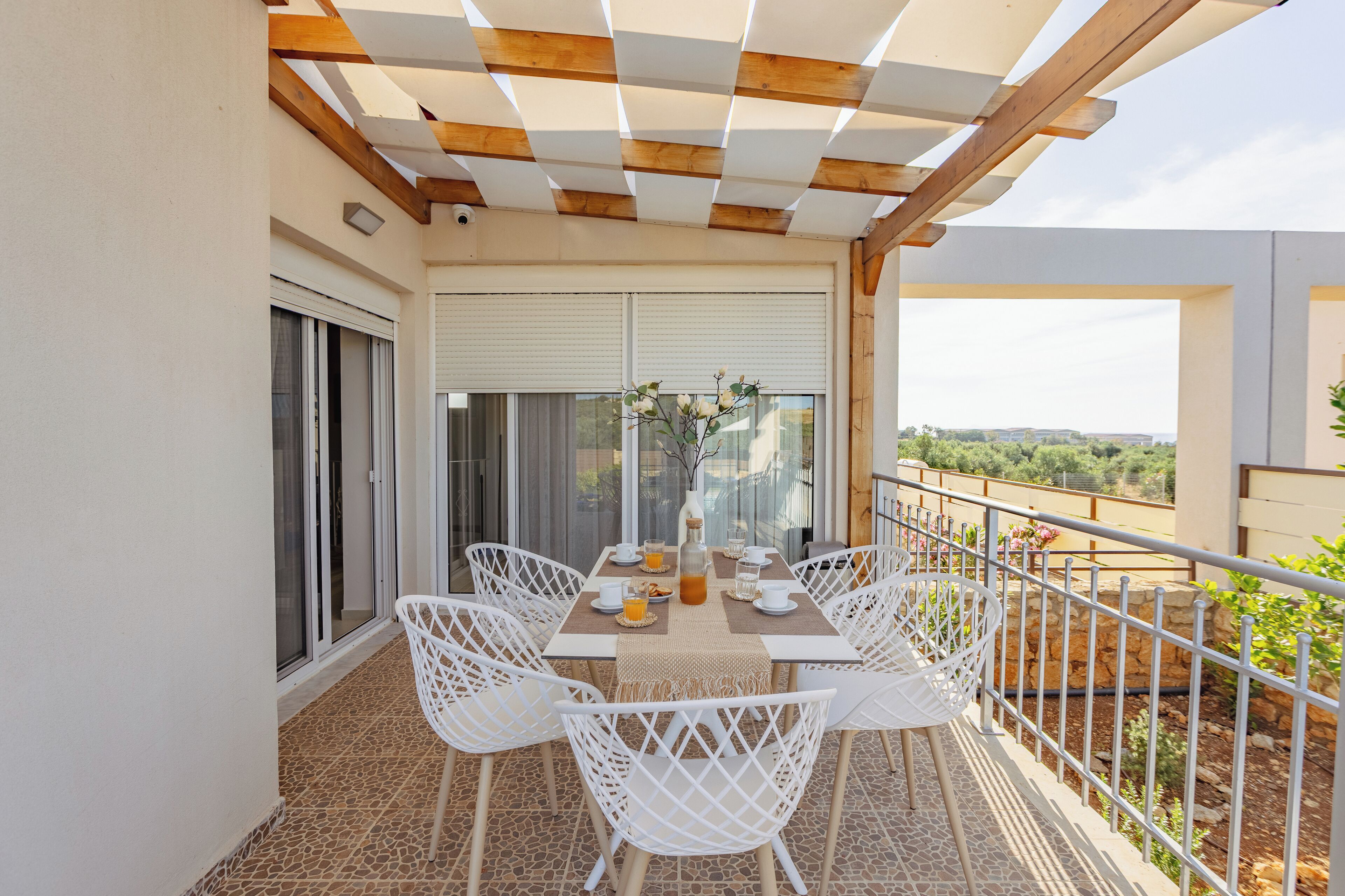 Ein Balkon mit Tisch für vier Personen, umgeben von weißen Stühlen, einer Pergola darüber und Blick auf die Landschaft.