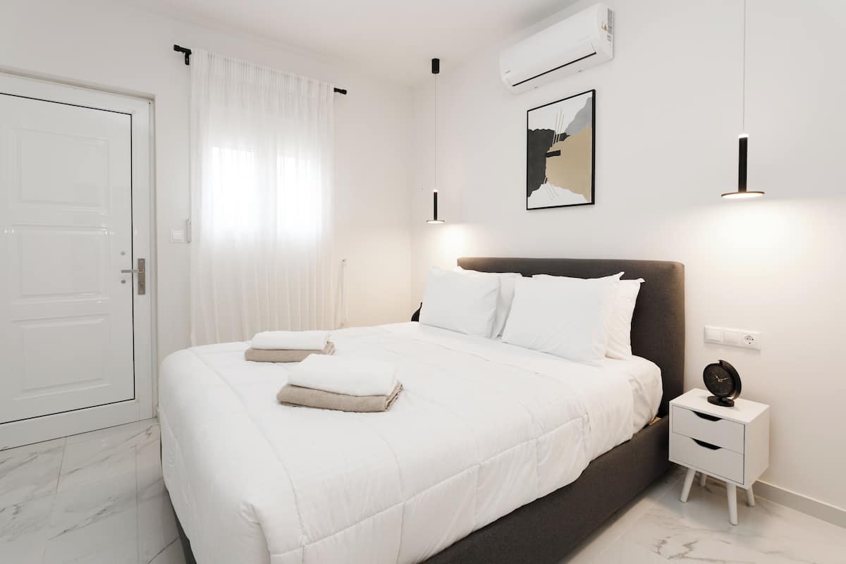 A modern bedroom with a large bed, white bedding, a nightstand with a clock, and a wall-mounted air conditioner.