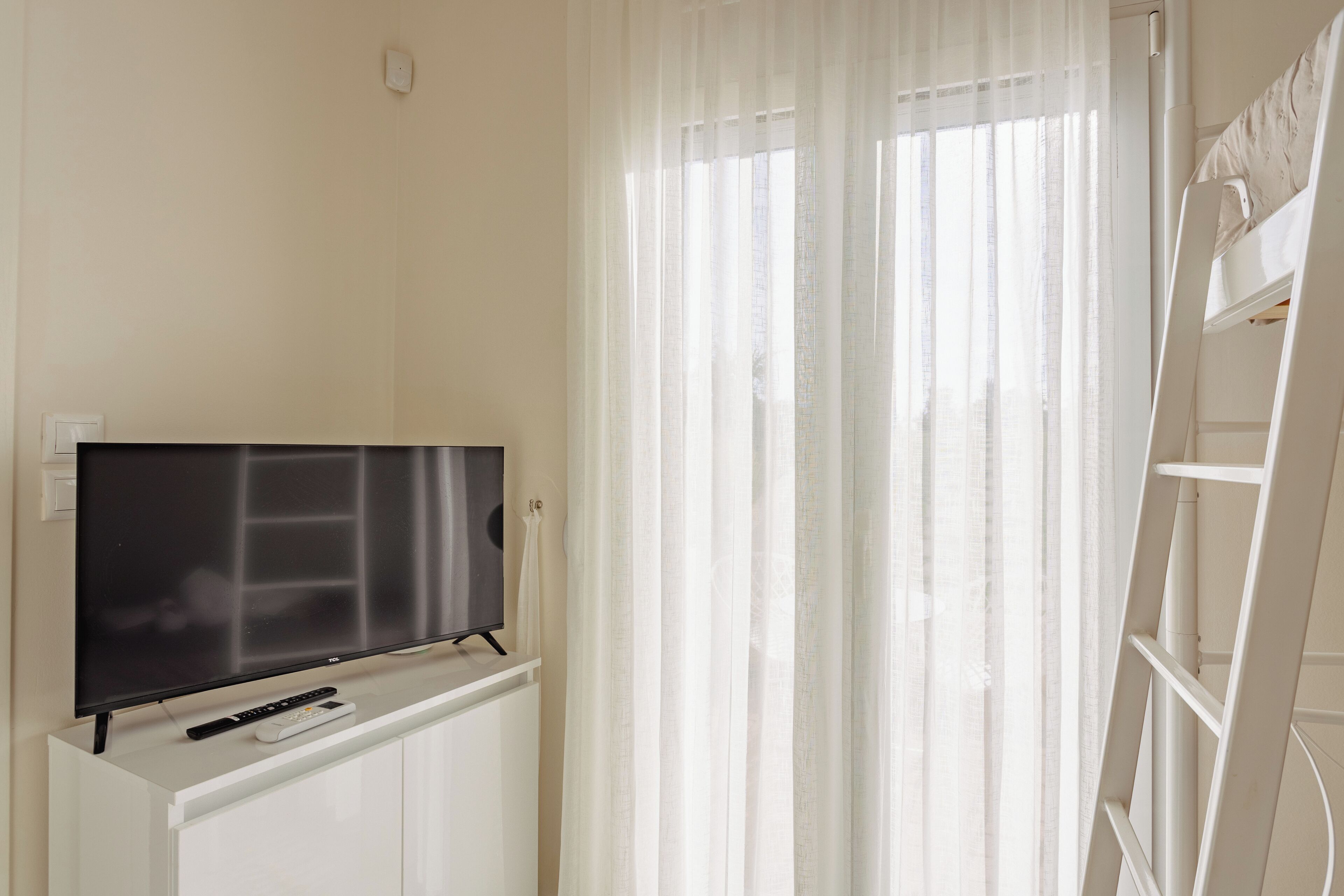 Una habitación con televisión sobre un mueble blanco, un control remoto y una ventana con cortinas de gasa.