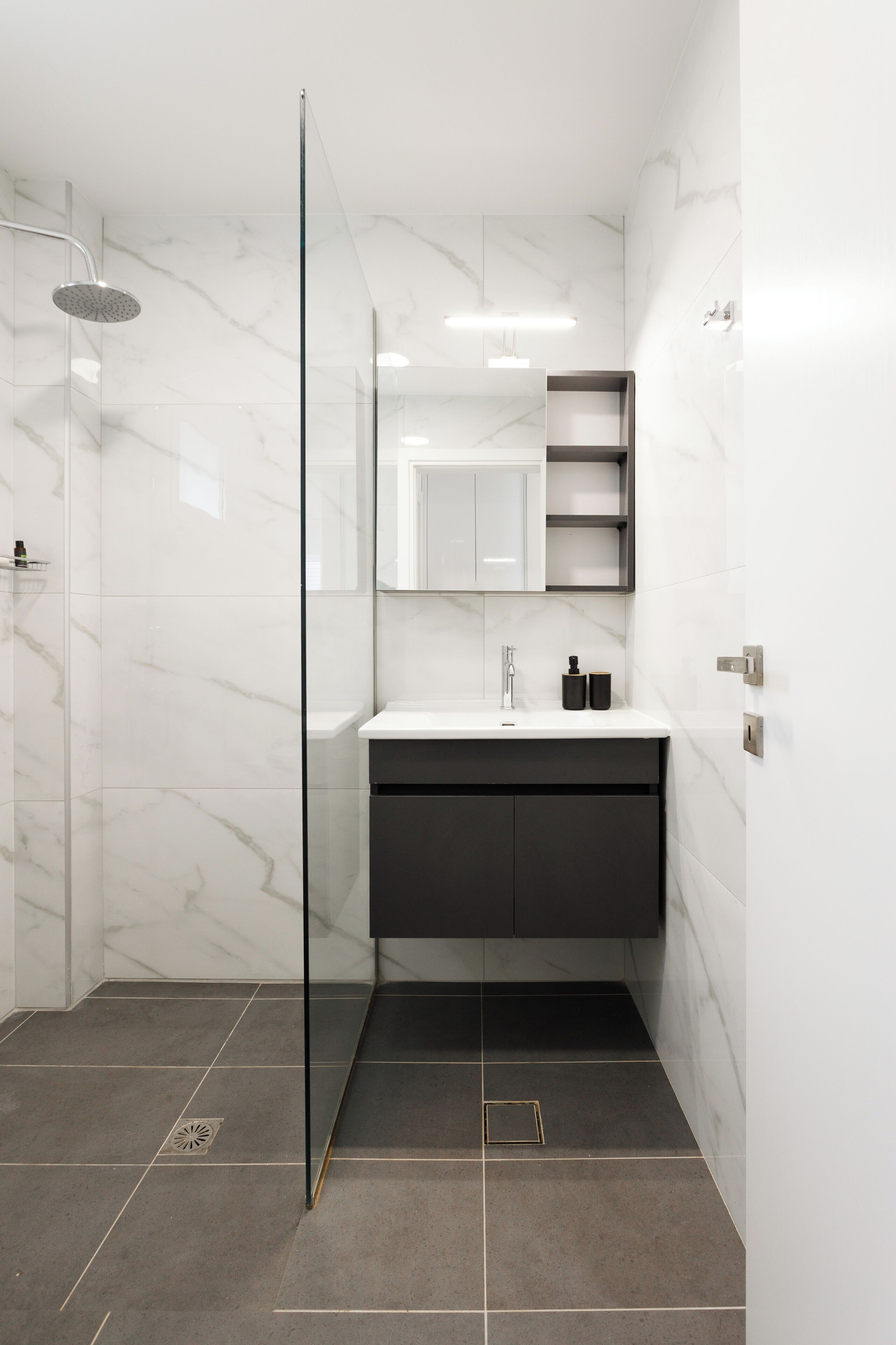 A modern bathroom with a glass shower enclosure, a white marble wall, a dark vanity, and a mirror above the sink.