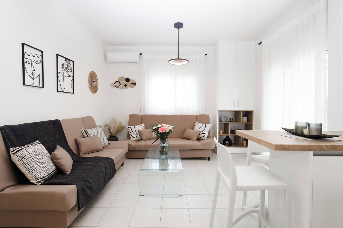 A modern living room with a beige sofa, a glass coffee table, and a white dining area.
