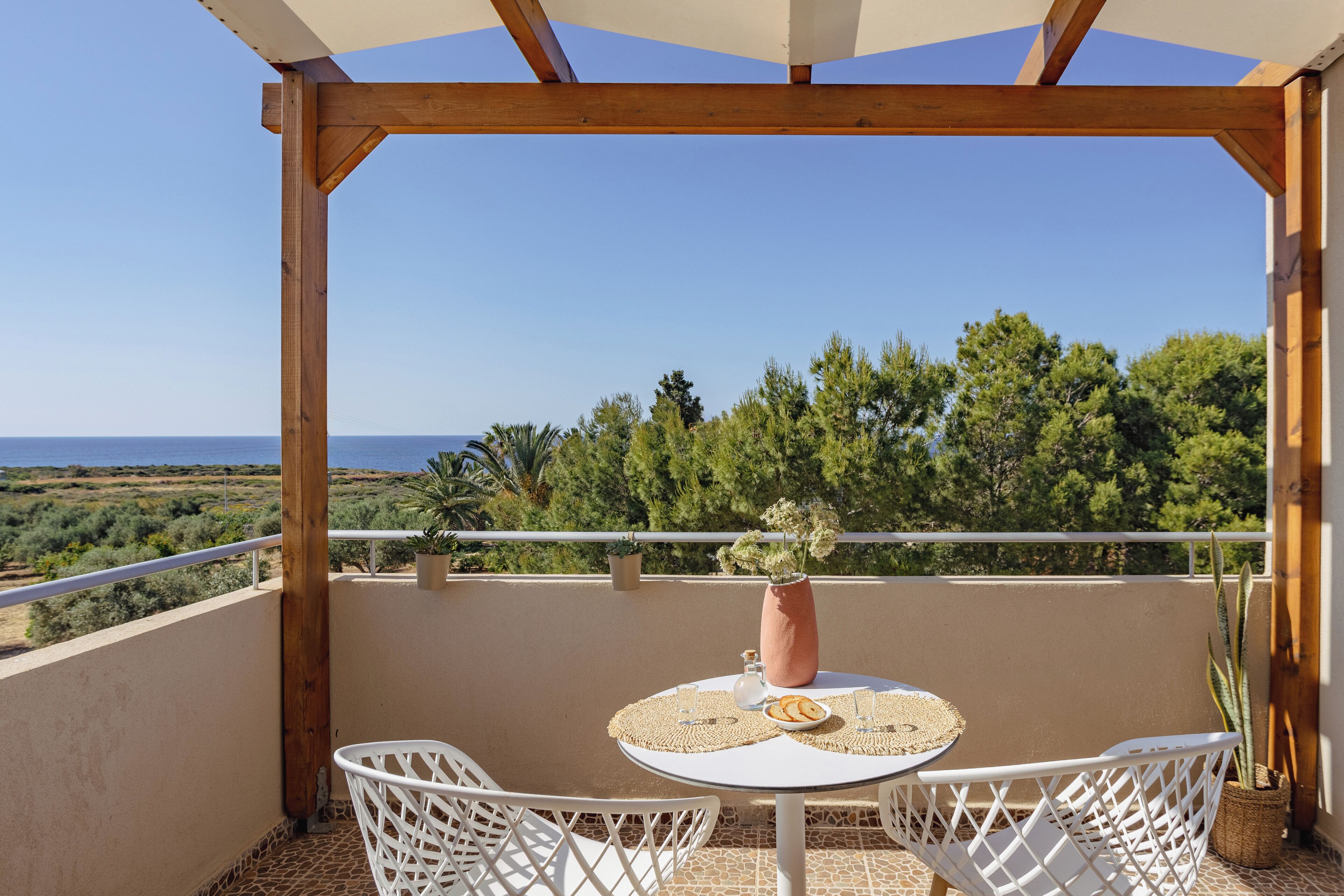 Un balcon avec une table et des chaises, un vase rempli de fleurs et une vue sur la mer et la verdure.