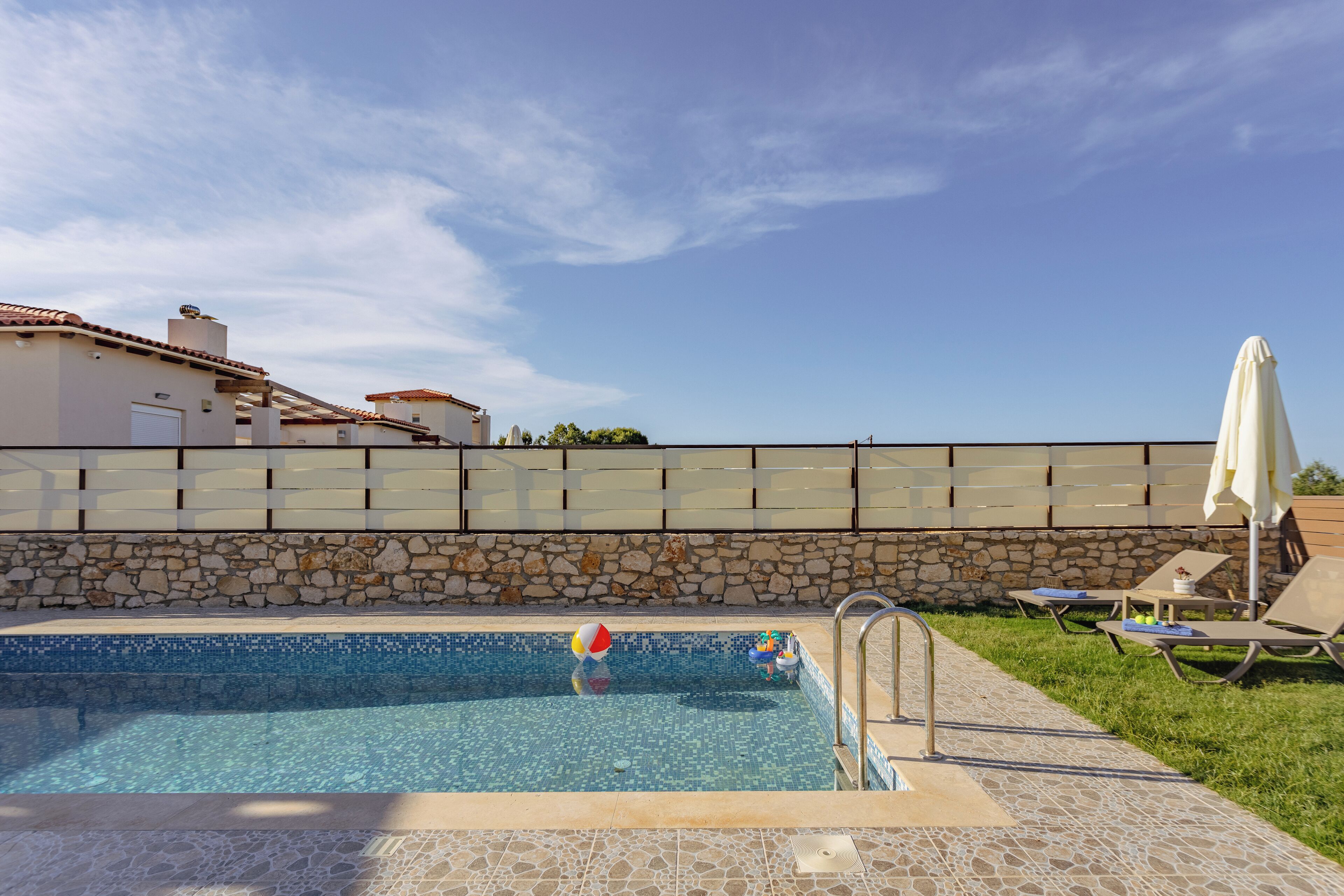 Una piscina con pared de piedra y solárium con baldosas, rodeada de sillones de descanso y sombrillas.