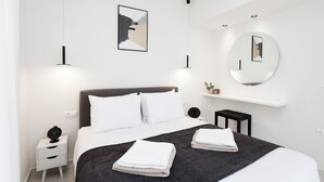A modern hotel room with a neatly made bed, a bedside table with a clock, a small side table, a round mirror, and a wall-mounted shelf with a plant. - Panor Villas (Rethymno)
