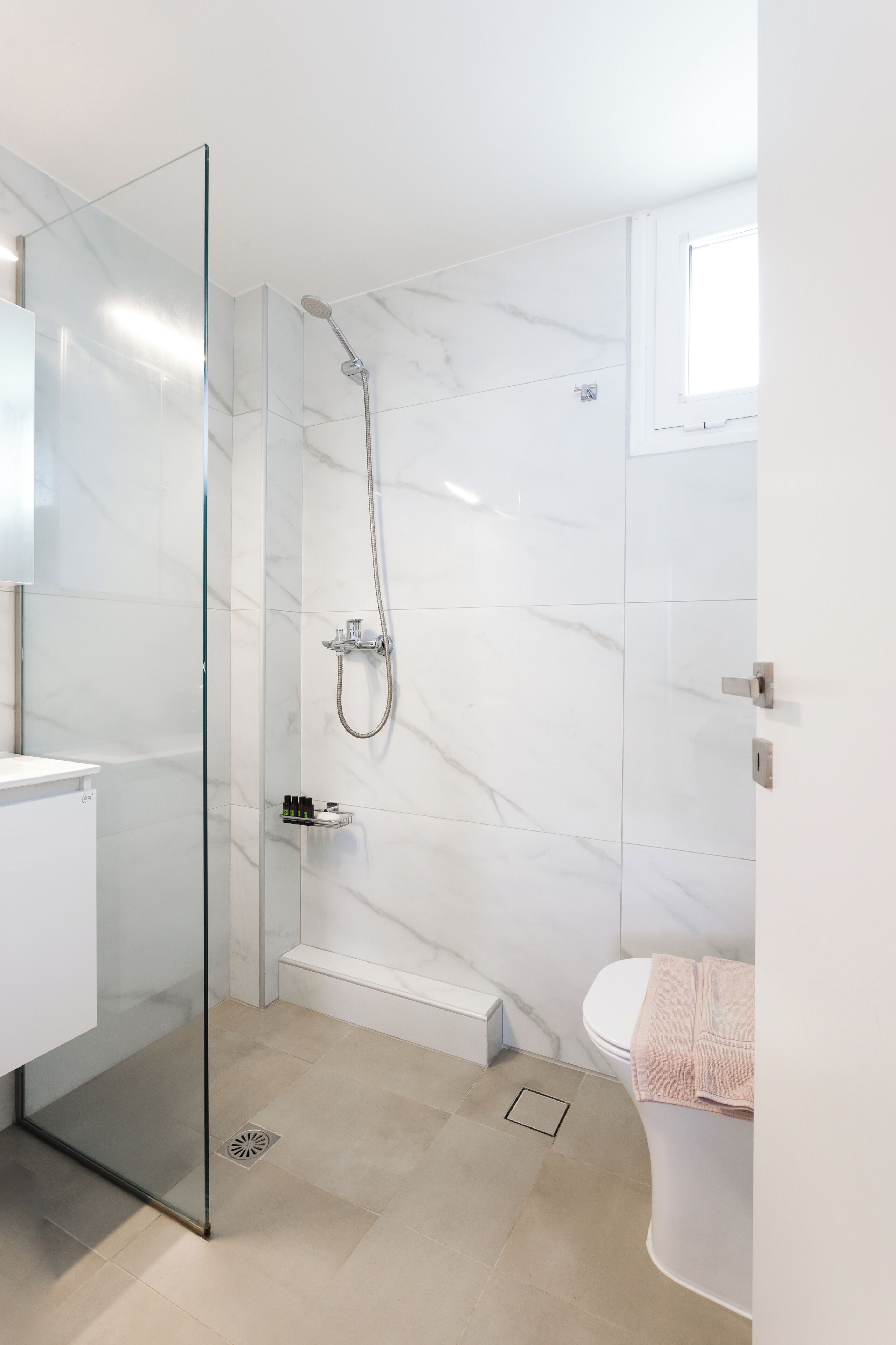 Une salle de bain moderne avec une douche à l’italienne en verre, des toilettes blanches et une petite fenêtre.