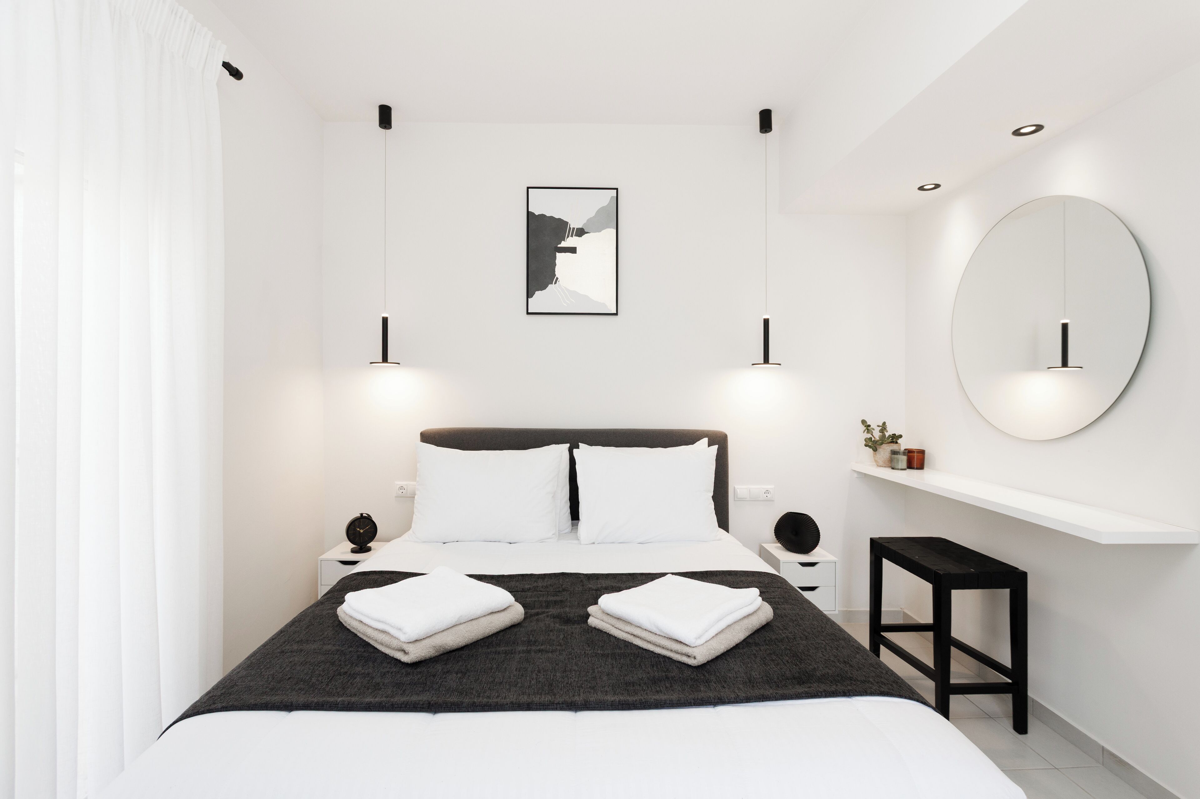 Une chambre à coucher moderne avec un grand lit, des tables de chevet, un miroir rond et une œuvre d’art encadrée accrochée au mur.