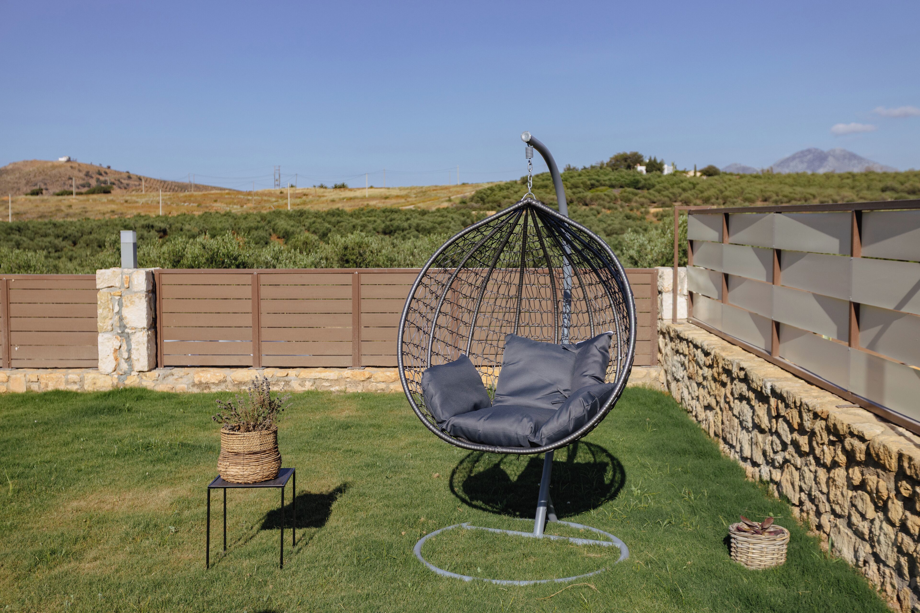 A hanging chair with cushions in a grassy outdoor area with a stone wall and a view of hills in the background.