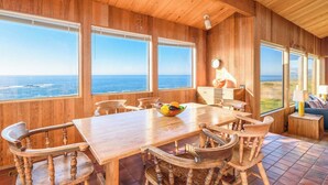 A wooden interior with a large table and chairs, large windows with ocean views, and a clock on the wall. - Sea Ranch Oceanfront 3BR | Hot Tub | Sleeps 6 (Sea Ranch)