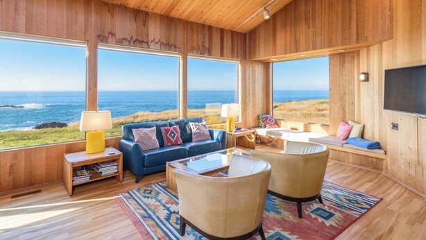 A spacious room with large windows offering ocean views, a blue sofa, two armchairs, a small table, and a TV mounted on the wall. - Sea Ranch Oceanfront 3BR | Hot Tub | Sleeps 6 (Sea Ranch)