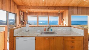 A kitchen with wooden cabinets, a stainless steel sink, and a dishwasher. There are large windows offering a view of the ocean. - Sea Ranch Oceanfront 3BR | Hot Tub | Sleeps 6 (Sea Ranch)