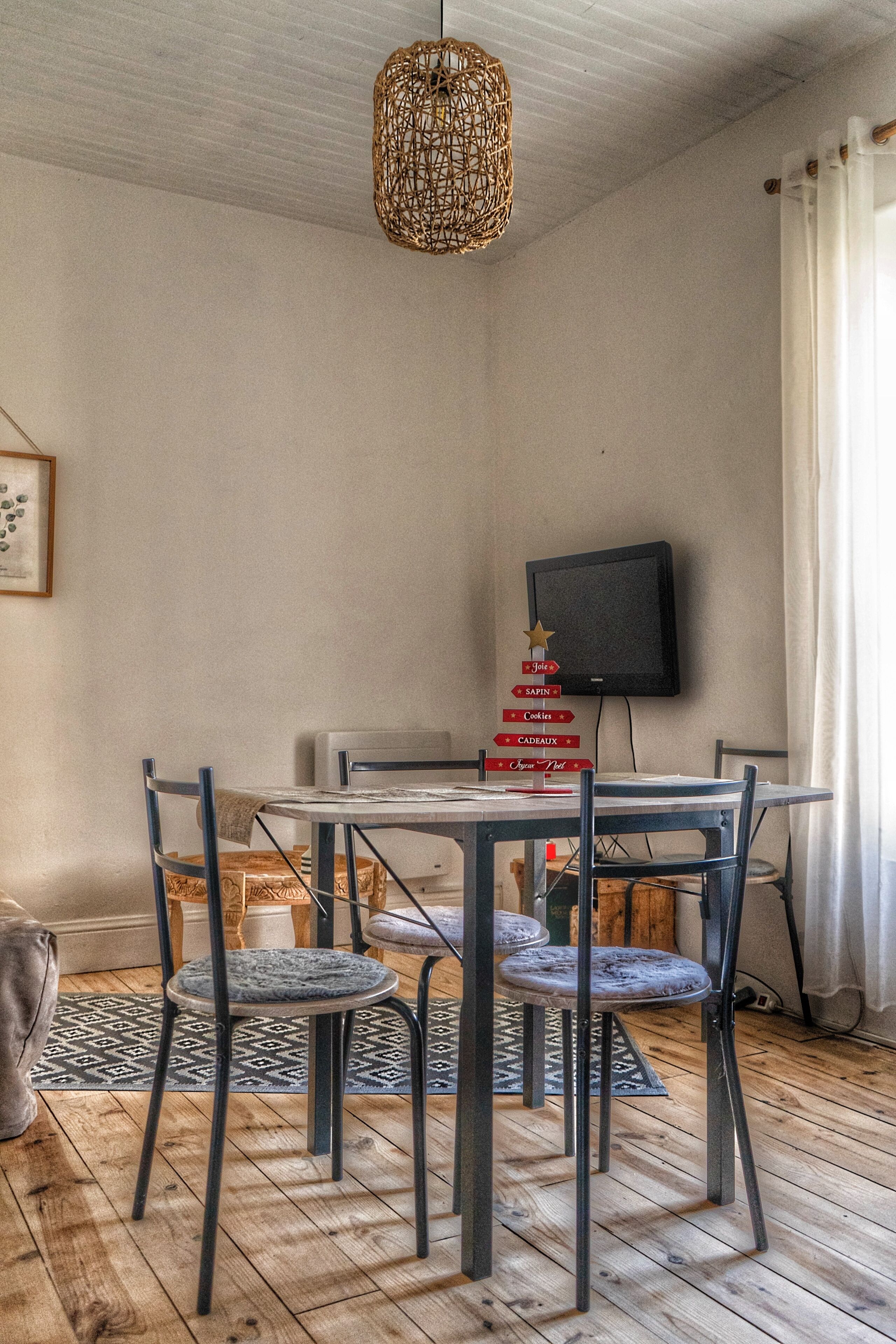 A dining area with a table and four chairs, a television mounted on the wall, and a hanging light fixture.