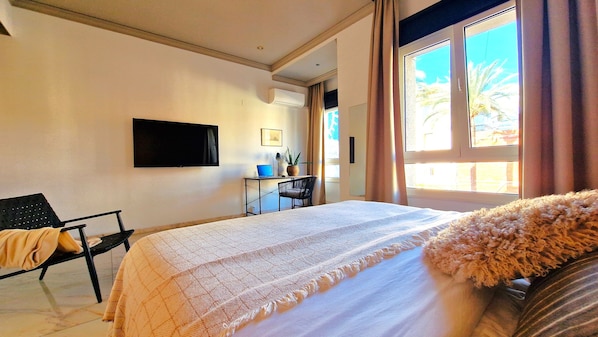 A hotel room with a bed, a desk with a laptop, a chair, a TV mounted on the wall, and a window with curtains.
