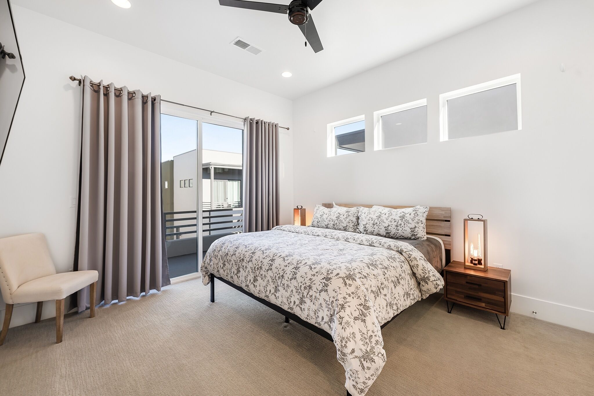 Un dormitorio con cama, una silla, una mesita de noche con lámpara, un ventilador de techo y una ventana con cortinas.