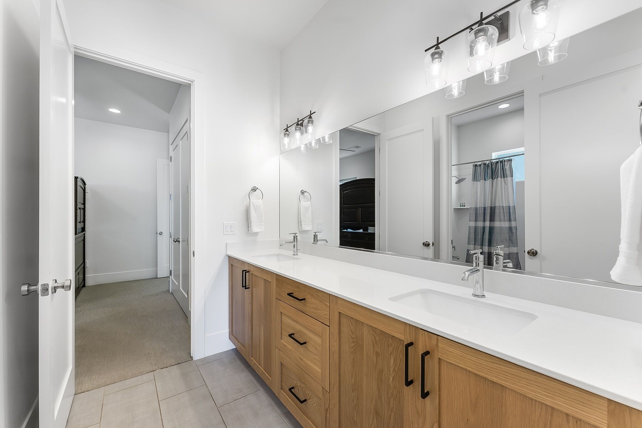 A modern bathroom with a large mirror, white countertop, wooden cabinets, and a shower area visible through an open door.
