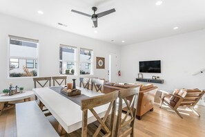 A modern living room with a dining area, wooden furniture, a ceiling fan, and a television on the wall.